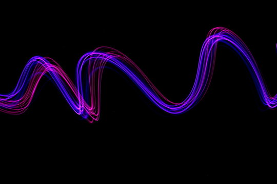 Long exposure photograph of neon pink and purple colour in an abstract swirl, parallel lines pattern against a black background. Light painting photography.