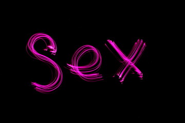 Long exposure photograph of the word 'sex' in pink neon colour in an abstract swirl pattern against a black background. Light painting photography.