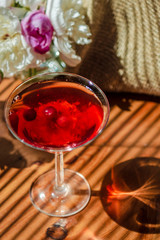 Solemn rustic still life with glass of red drink with autumn berries and white flowers on a wooden table and textile in the diagonal striped shadow of the blinds in bright sunny day. Rich colors