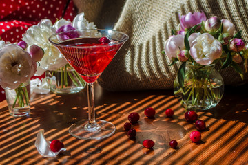 Rustic still life with glass of red drink with autumn berries surrounded by white flowers on a textile background on a wooden table in the diagonal striped shadow of the blinds on a bright sunny day
