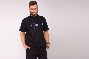 Fototapeta premium young stylish nurse male in black medical costume with stethoscope on his neck is standing with hands in pockets and smiling on the white wall background. medical concept. free space on right side