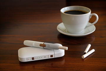 Heating tobacco system and a cup of coffee on a brown table, e-cigarette with tobacco sticks that...