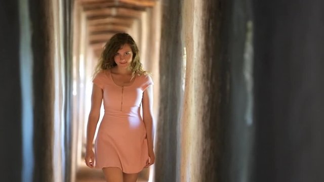 Young girl happy smiling woman in light pink dress walking in slow motion in fortress tunnel passage in Italy during summer with walls and windows in Castiglione del Lago