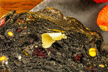 Obraz premium Détail sur une tranche de pain noir au charbon fruits secs et cranberry sur un plat en ardoise avec des fruits frais fraises et une noisette de beurre