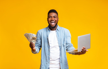 Overjoyed black guy holding cash and laptop over yellow background
