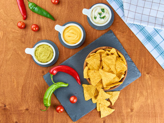 Nachos - yellow corn chips with various sauces in bowls: guacamole, cheese sauce, white sauce, on a wooden table. Mexican food concept. The view from the top.