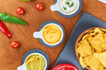 Nachos - yellow corn chips with various sauces in bowls: guacamole, cheese sauce, white sauce, on a wooden table. Mexican food concept. The view from the top.