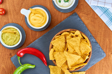 Nachos - yellow corn chips with various sauces in bowls: guacamole, cheese sauce, white sauce, on a wooden table. Mexican food concept. The view from the top.