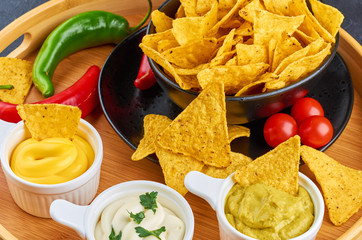 Nachos - yellow corn chips with various sauces in bowls: guacamole, cheese sauce, white sauce, on dark background. Mexican food concept.
