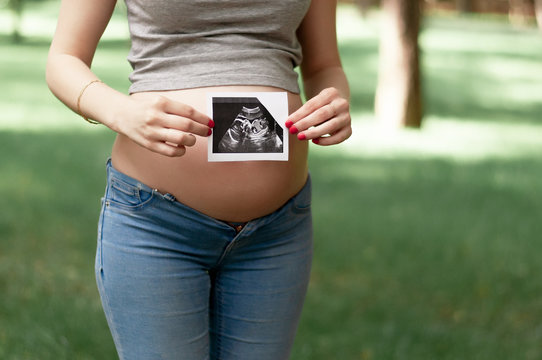 Happy young adult pregnant girl holds in her hands a snapshot of an ultrasonic study of a toddler walking in a park. Second trimester.