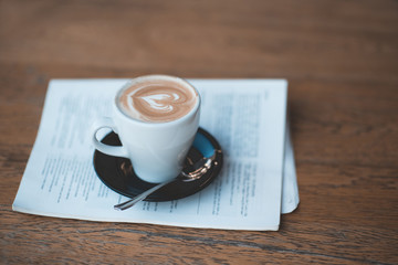 Cute cup of coffee with heart on milk froth staying on open magazine on wooden tale in kitchen close up. Breakfast time. Good morning.