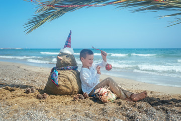 Little boy and Snowmen at tropical ocean beach in santa hat. New Years and Christmas holiday in hot countries concept.