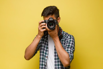 Handsome and confident indian man photographer with a large professional camera taking pictures photo shooting on the on the yellow background. - obrazy, fototapety, plakaty