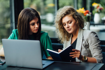 Two young business women having lunch break together in a coffee shop terrace, using laptop and notebook.
