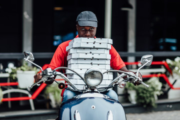 Young African courier poses on fast motorbike, carries rucksack, wears helmet, holding cardboard, looks aside with pleased expression. People, technology, transport.