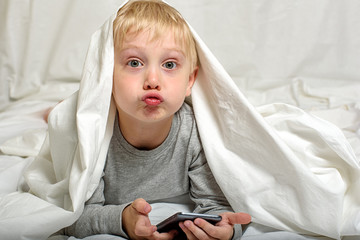 Little blond boy grimaces with a smartphone in his hands. Lying in bed and hiding under the covers. Gadget Leisure