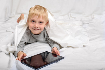 Smiling little blond boy with a tablet. Lying in bed and hiding under the covers. Gadget Leisure
