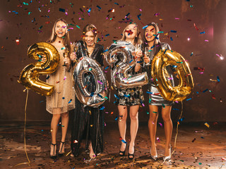 Fototapeta na wymiar Beautiful Women Celebrating New Year. Happy Gorgeous Girls In Stylish Sexy Party Dresses Holding Gold and Silver 2020 Balloons, Having Fun At New Year's Eve Party. Holiday Celebration.Raising hands