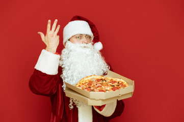 Portrait of positive Santa Claus in glasses isolated with pizza box in hands on red background, looking into camera. Man in red santa suit posing with pizza on red background. Christmas