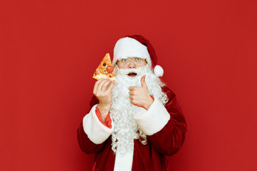 Happy Santa Claus in a red suit stands on a red background with a piece of pizza in his hand, rejoices and shows his thumb up. Santa loves to eat pizza. Pizza for Christmas. New year with pizza.