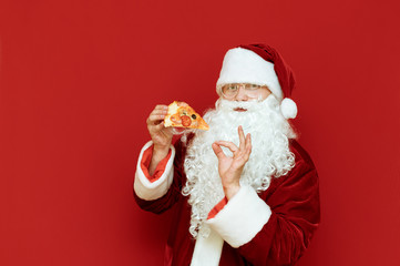 Man in a Santa costume eats a pizza. Pizza for Christmas . Handsome Santa with a slice of pizza in her hand stands on a red background, looks in camera and shows her thumbs sign OK. Copyspace