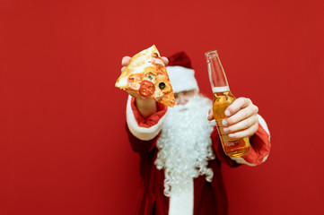 Santa Claus shows in the camera a bottle of beer and a slice of pizza on a red background. Pizza and bottle in santa's hands isolated on red background. Christmas concept.