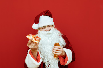 Cheerful santa portrait with fast food meal, isolated. Funny Santa Claus in a classic costume stands on a red background with a slice of pizza and a bottle of beer, laughs with his eyes closed