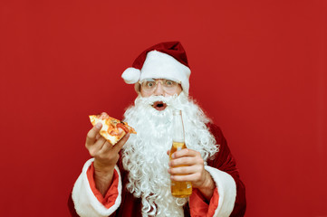 Surprised Santa Claus with piece of pizza and bottle of beer in hand stands on red background, looks into camera with shocked face, isolated. Funny Santa eats junk food and drinks alcohol.