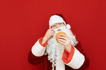 Santa Claus in red costume and beard isolated on red background, holds a tasty burger in his hands and is about to eat it. Funny Santa eating fast food on red wall background. Fast food and Christmas.