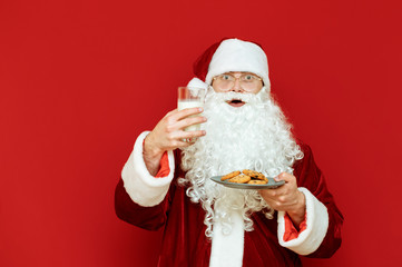 Portrait of surprised Santa Claus with glass of young and plate of cookies in hands, isolated on red background, with shocked face looks into camera. Christmas and New Year concept. Tastes for Santa