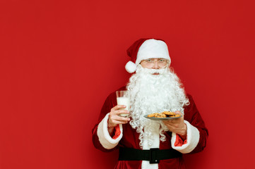 Cute Santa Claus stands on a red background with a glass of milk and a plate of chocolate cookies, looks into the camera and smiles. Christmas and New Year concept. Isolated. Copyspace