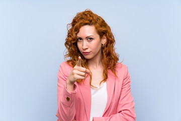 Redhead woman in suit over isolated blue wall frustrated and pointing to the front