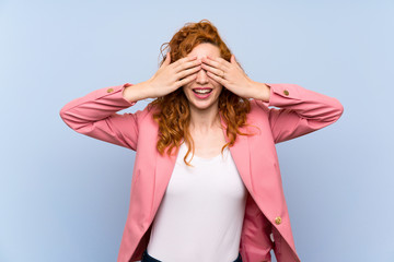 Redhead woman in suit over isolated blue wall covering eyes by hands