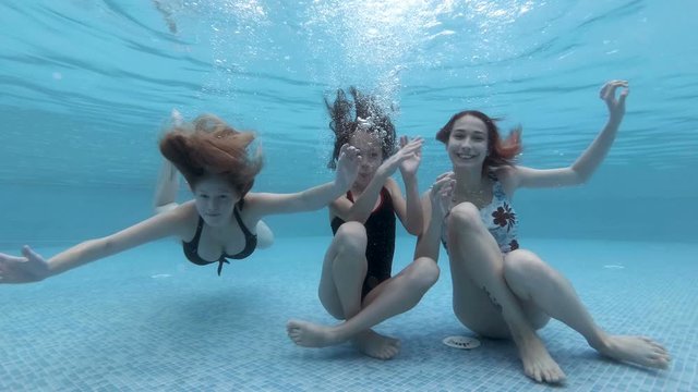 Three young girls of different ages swim and play underwater in the pool. They dive and sit at the bottom of the pool, look at the camera, wave and smile. Portrait. Slow motion. 4K.