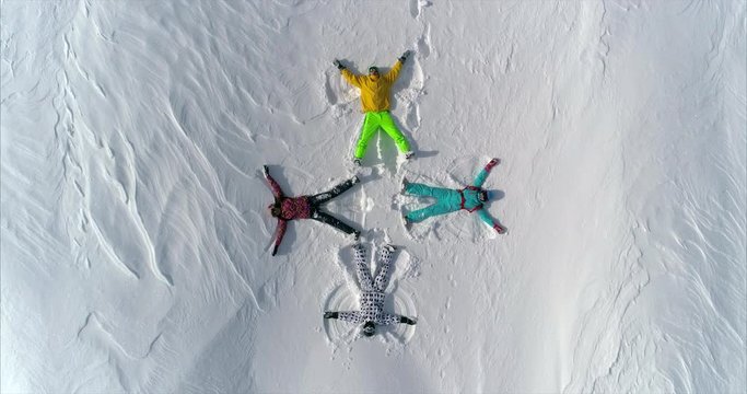 AERIAL TOP UP: a group of young people in bright ski suits depict snow angels lying on the fresh snow in the form of a cross. four winter angels happy and energetic with splashes of snow. SLOW MOTION