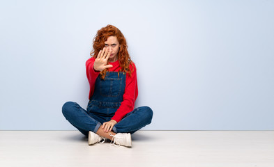 Redhead woman with overalls sitting on the floor nervous stretching hands to the front