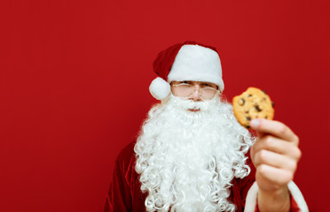 Serious man in Santa Claus costume stands on red background with cookies in hand, looks into camera and shows cookies. Focus on Santa Claus with cookies in hand. Close up Christmas portrait.