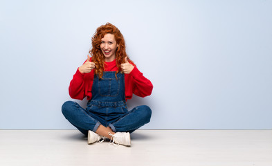 Redhead woman with overalls sitting on the floor with thumbs up gesture and smiling