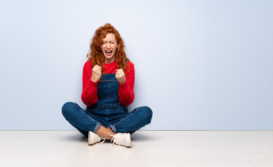 Redhead woman with overalls sitting on the floor frustrated by a bad situation