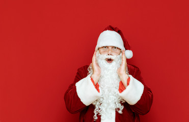 Fototapeta na wymiar Astonished Santa Claus stands against a red wall and looks into the camera. Portrait of shocked man in santa costume isolated. Santa was surprised at the discounts for Christmas. Copyspace