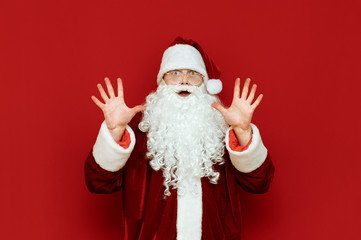Portrait of a frightened Santa Claus stands on a red background and looks shockingly at the camera, raising his hands up in fear. Surprised Santa is scared. Isolated. Copyspace