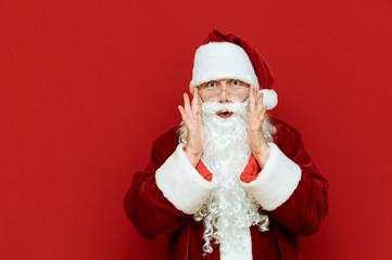 Portrait of surprised Santa Claus on red background, corrects glasses and looks shockingly at camera. Isolated. Emotional Santa Claus is amazed. Christmas and New Year. Copyspace