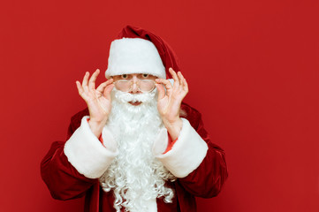 Surprised Santa Claus looks carefully at the camera isolated on red background. The man in Santa's suit with a shocked face looks into the camera, Santa can't believe it. Christmas discounts. Sale