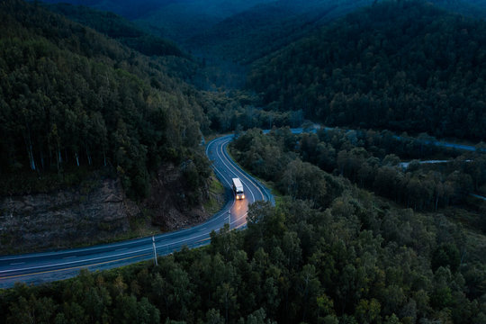 Aerial view of a sharp turn on a mountain road among green forest trees. Semi truck with cargo trailer and bright headlights on a dark highway. P-258 road near Baikal shore in Siberia, Russia