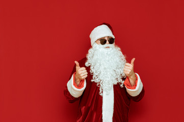 Fashionable Santa Claus in black sunglasses isolated on red background, shows thumbs up and looks into the camera. Studio photo of Santa showing gesture like. Christmas and New Year concept.