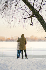 Theme love and active rest on rustic nature. Young couple Caucasian guy and girl stand back in full growth holding hands in winter on snow pier overlooking frozen lake beautiful view of horizon