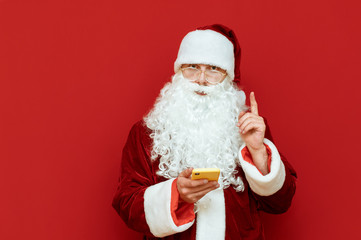 Portrait of Santa Claus on red background with smartphone in hand stands on red background, looks into camera with serious face and raised forefinger. Christmas concept. Copyspace