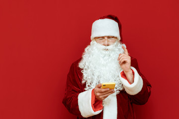 Concentrated Santa Claus uses a smartphone and raises his finger, he has an idea, stands on a red background and looks at a smartphone screen. Santa Claus and technology. Christmas concept. Copyspace