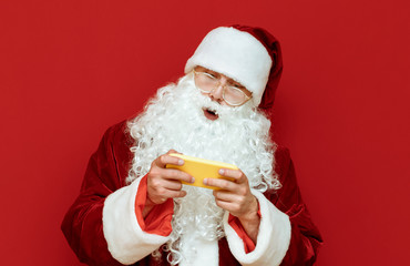 Fototapeta na wymiar Santa Claus gamer plays video games on smartphone, looks surprised at screen isolated on red background. Portrait of santa gamer. Christmas gaming.