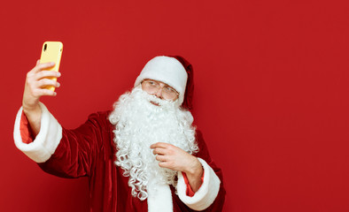  Santa Claus stands on a red background with a smartphone in his hand and takes a selfie, looks into the camera and smiles. Santa Claus and the modern techie, uses a gadget.  Christmas selfie.